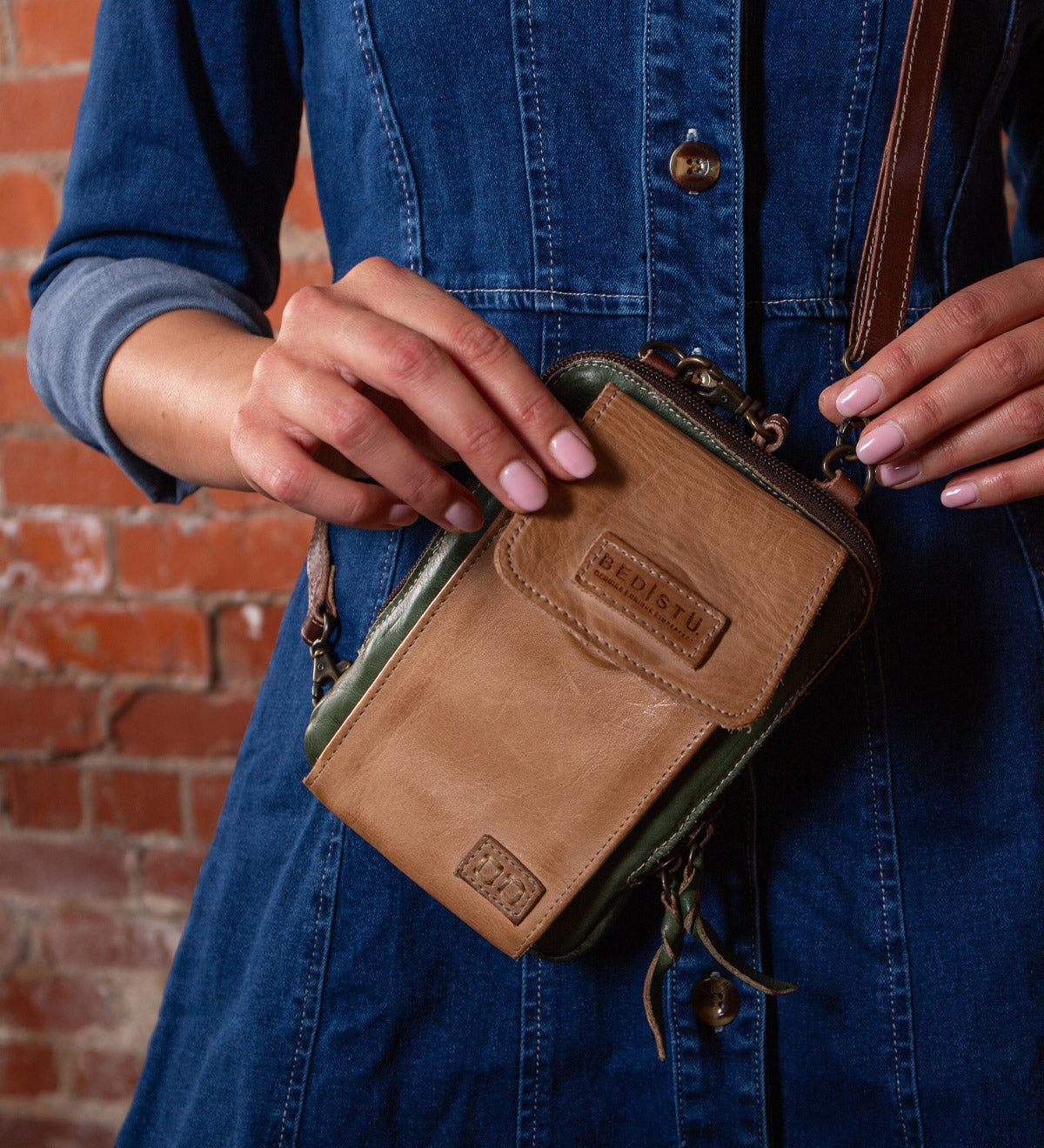 A person in a denim dress holds the Bed Stu Alelike, a premium leather compact crossbody phone holder, in front of a brick wall.