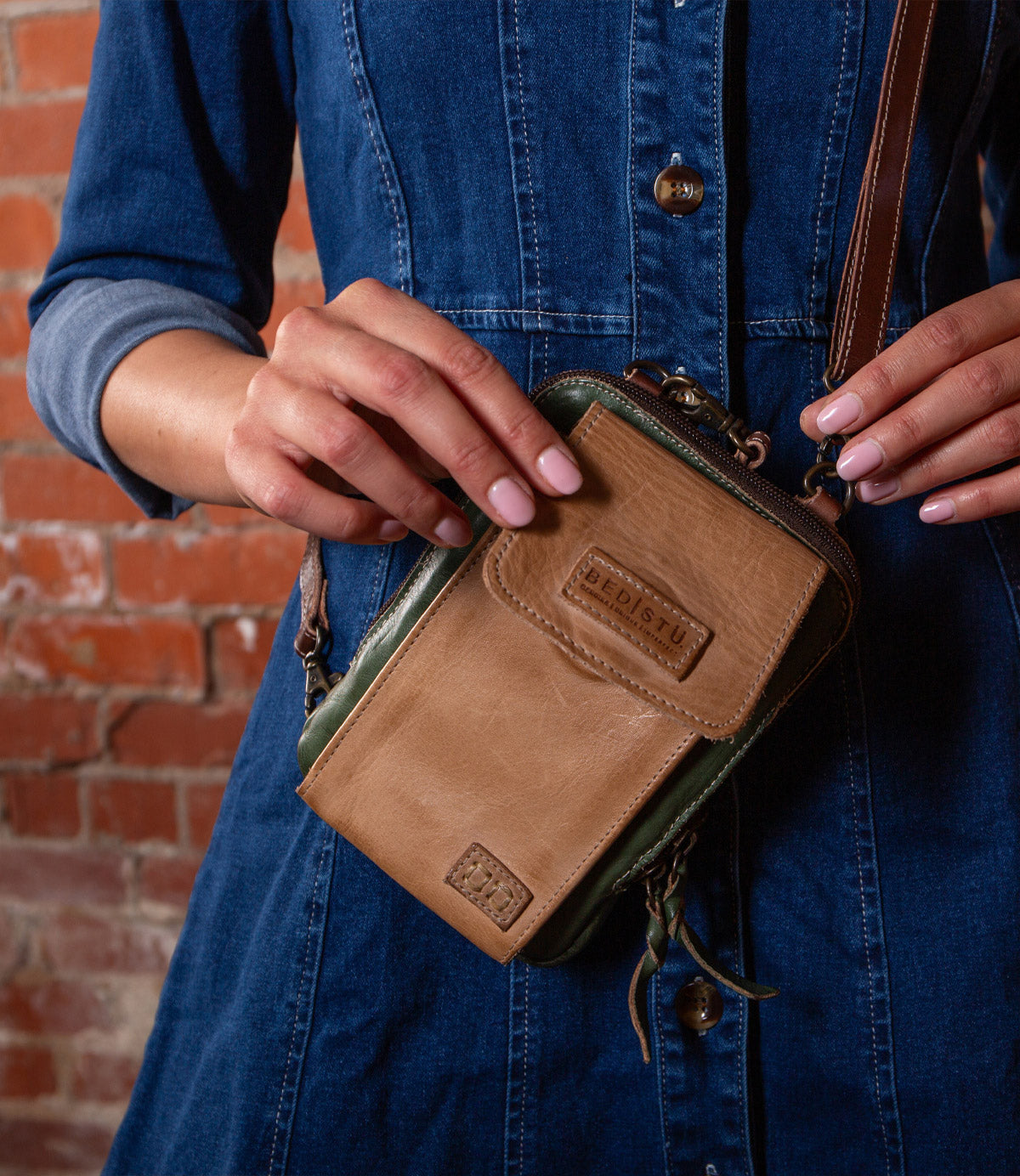 A person in a denim dress holds the Bed Stu Alelike, a premium leather compact crossbody phone holder, in front of a brick wall.