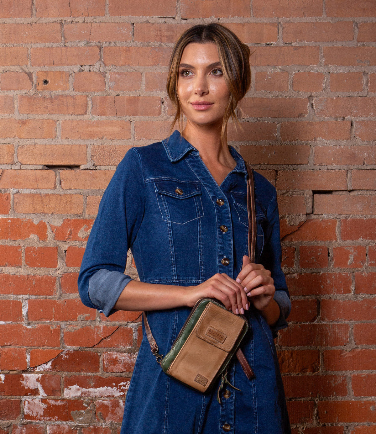 A woman in a blue denim dress stands by a brick wall, holding the Alelike crossbody phone holder by Bed Stu, crafted from tan and green premium leather.