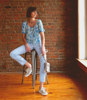 A woman is sitting on a stool in front of a brick wall, with a Bed Stu premium leather Alelike crossbody phone holder.