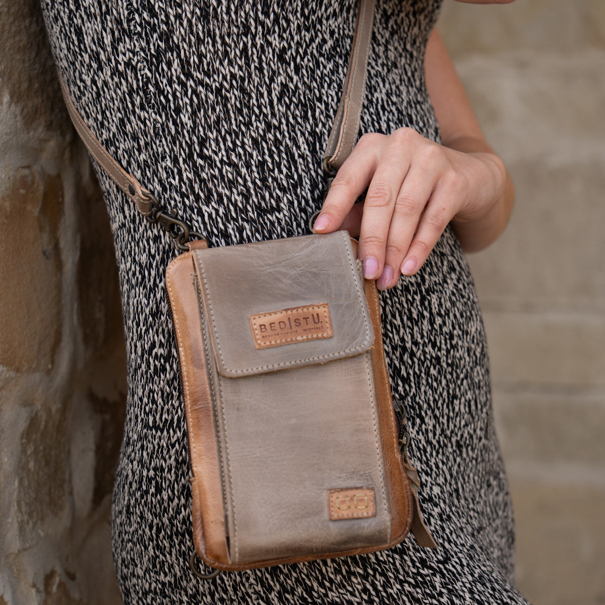 A person in a patterned dress holds the compact brown Alelike crossbody bag by Bed Stu against a stone wall.