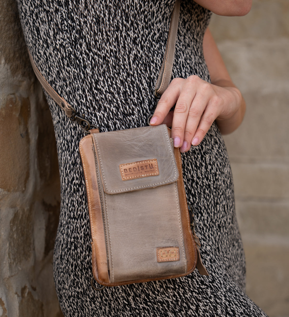 A person in a patterned dress holds the compact brown Alelike crossbody bag by Bed Stu against a stone wall.