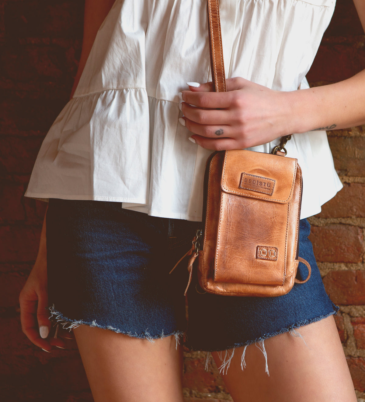 A woman in denim shorts holding a Bed Stu Alelike premium leather crossbody bag.