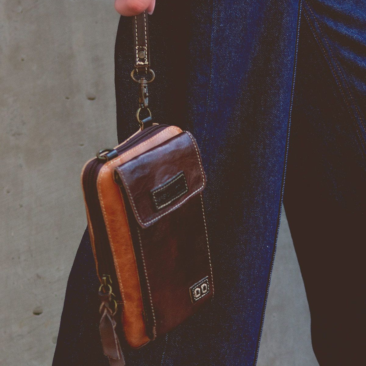 A person holds the Alelike, a small compact crossbody phone holder made of premium leather by Bed Stu, by a wrist strap, wearing dark blue jeans against a plain background.