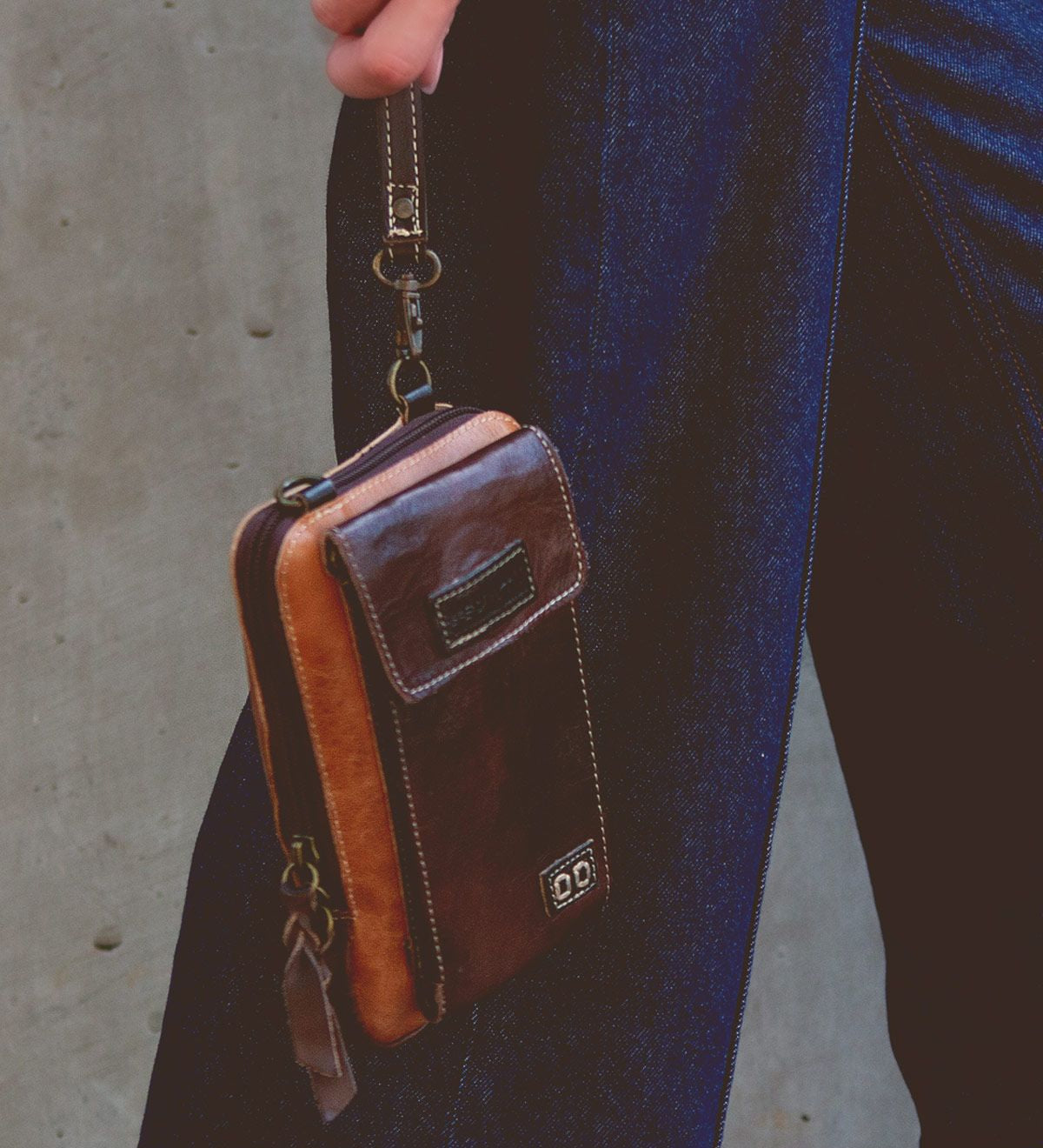 A person holds the Alelike, a small compact crossbody phone holder made of premium leather by Bed Stu, by a wrist strap, wearing dark blue jeans against a plain background.