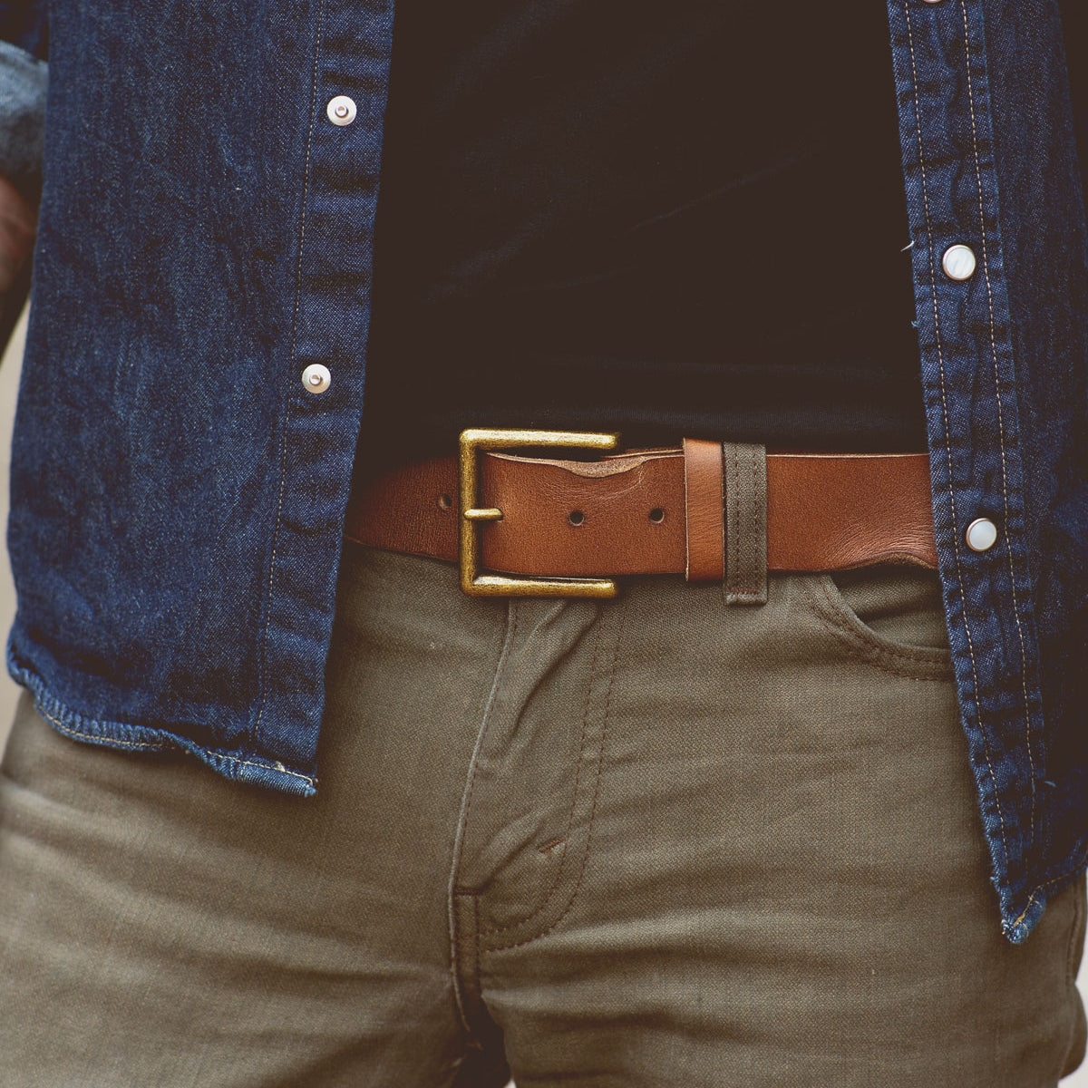 Close-up of a person wearing a blue denim shirt, olive-green pants, and an unisex brown leather belt with a removable metal buckle (Alex by Bed Stu), with one hand in a pocket.