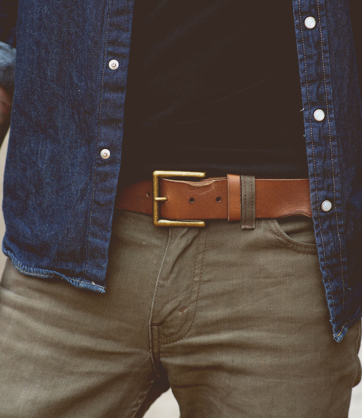 Close-up of a person wearing a blue denim shirt, olive-green pants, and an unisex brown leather belt with a removable metal buckle (Alex by Bed Stu), with one hand in a pocket.