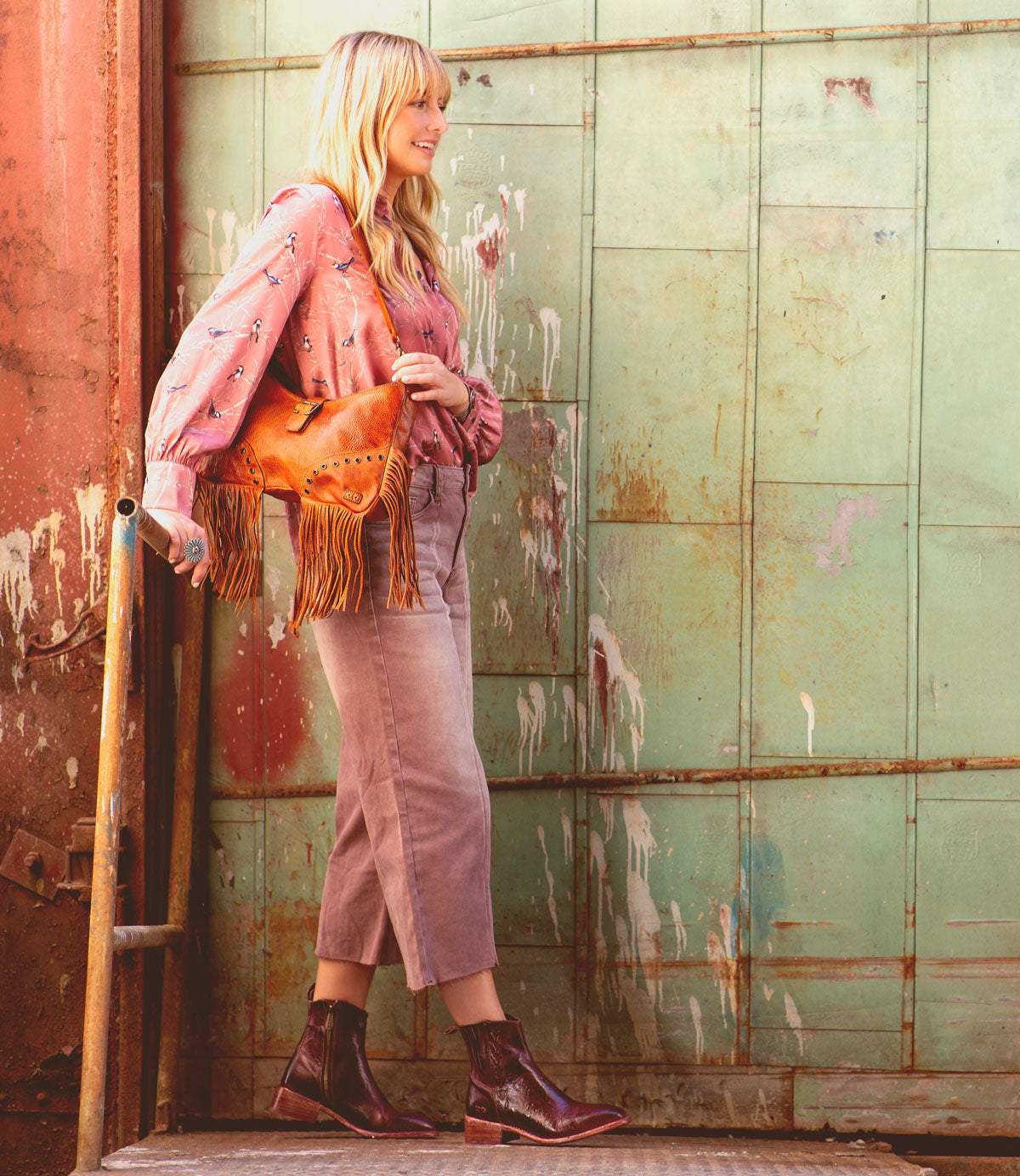 A Western-themed blonde woman is casually leaning against a city wall, effortlessly holding the Bed Stu Alina leather bag.