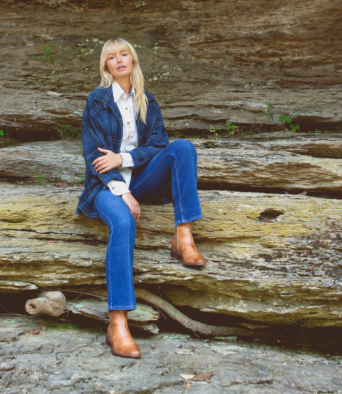 A blonde woman sitting on a rock wearing Bed Stu Alina leather ankle boots with western charm.