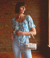 A woman in blue jeans and a floral top holding a sleek Bed Stu Amina II clutch.