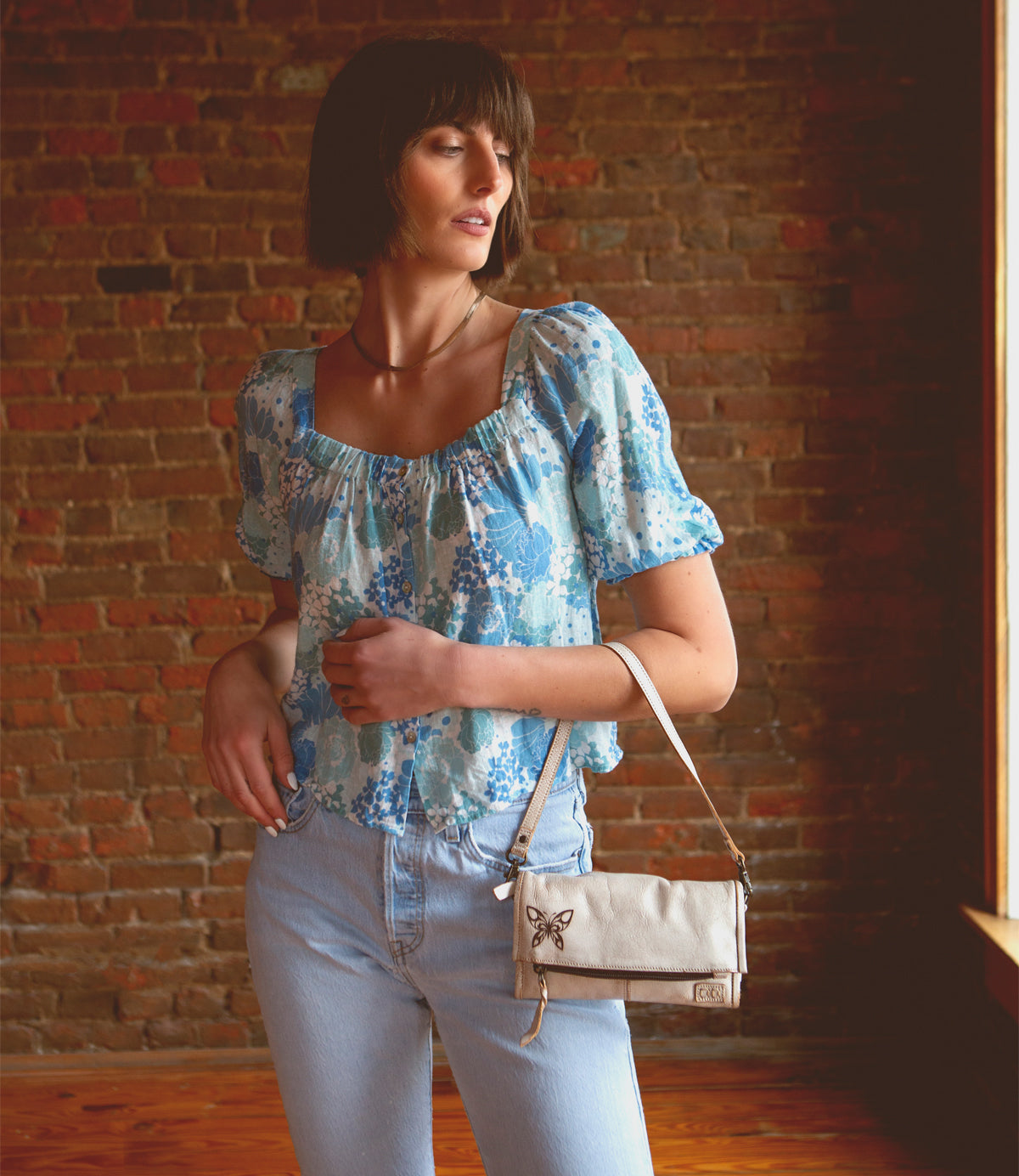 A woman in blue jeans and a floral top holding a sleek Bed Stu Amina II clutch.