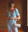 A woman in blue jeans and a floral top holding a sleek Bed Stu Amina II clutch.