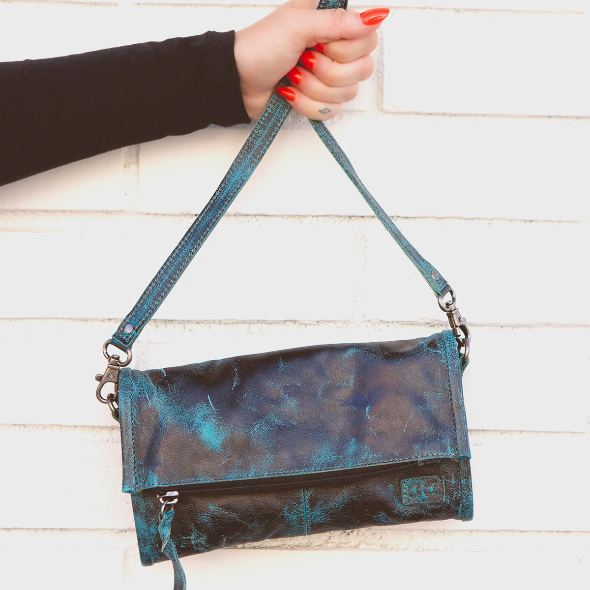 A hand with red-painted nails is holding the Amina blue and black leather clutch by its strap against a white brick wall from the Bed Stu brand.
