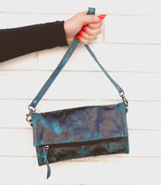 A hand with red-painted nails is holding the Amina blue and black leather clutch by its strap against a white brick wall from the Bed Stu brand.
