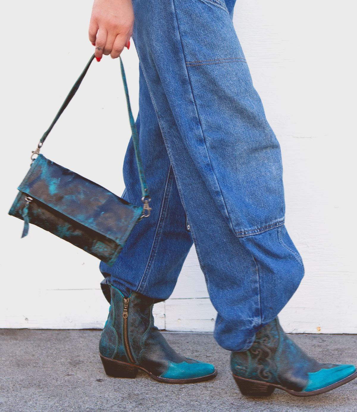 Person wearing blue denim pants and blue cowboy boots, holding the Bed Stu Amina crossbody handbag with a blue and green pattern while walking.
