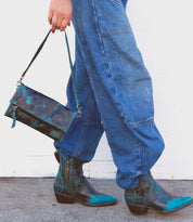 Person wearing blue denim pants and blue cowboy boots, holding the Bed Stu Amina crossbody handbag with a blue and green pattern while walking.