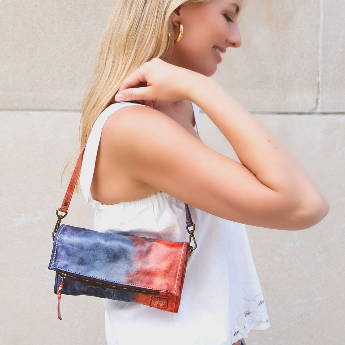 A woman with blonde hair, wearing a white sleeveless top, stands outdoors holding a Bed Stu Amina compact crossbody handbag over her shoulder, exuding versatile sophistication.