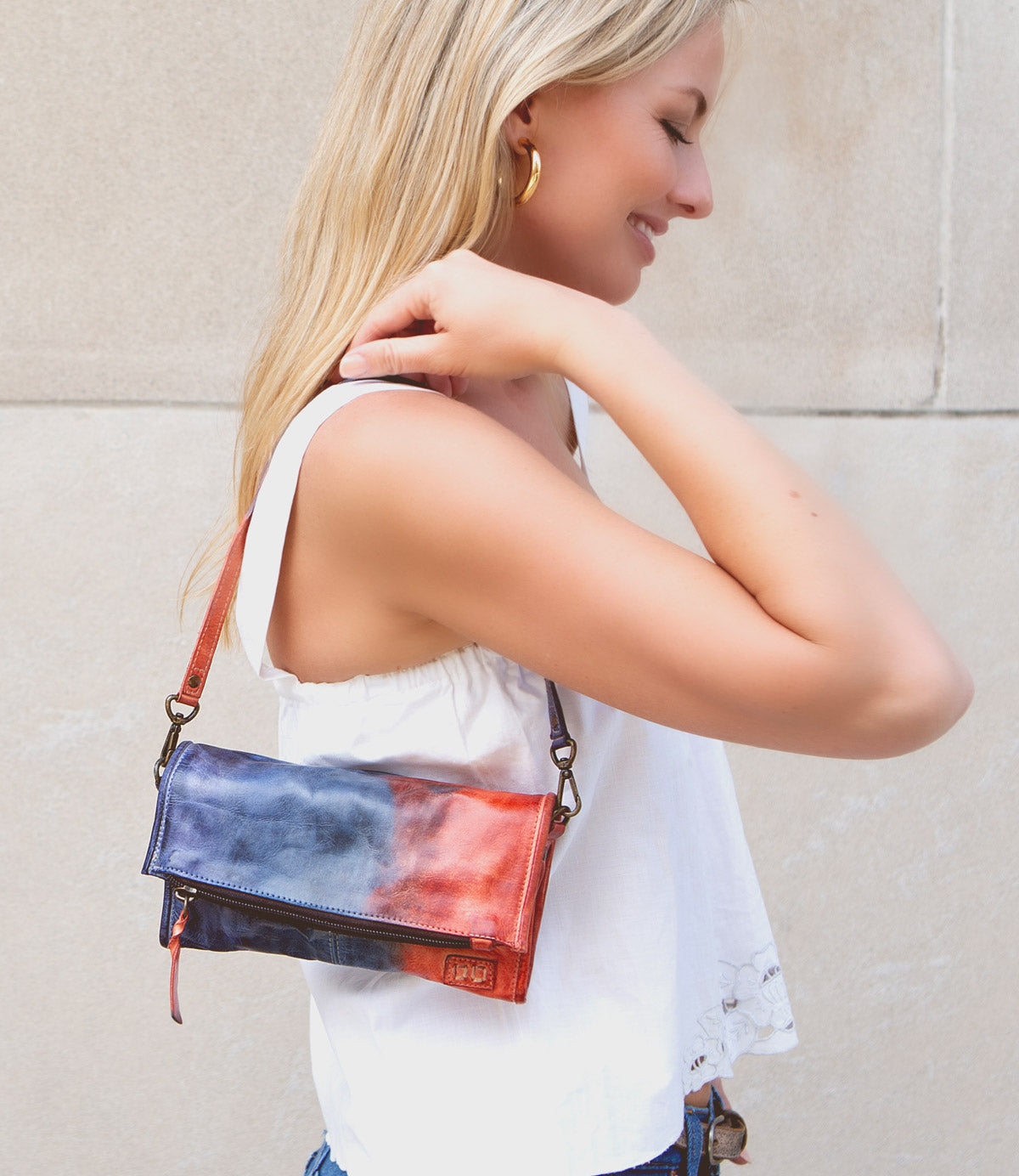 A woman with blonde hair, wearing a white sleeveless top, stands outdoors holding a Bed Stu Amina compact crossbody handbag over her shoulder, exuding versatile sophistication.