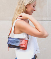 A woman with blonde hair, wearing a white sleeveless top, stands outdoors holding a Bed Stu Amina compact crossbody handbag over her shoulder, exuding versatile sophistication.