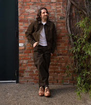 A man wearing the Anchiano brown jacket and pants by Bed Stu, a white t-shirt, and oxford lace-up tan shoes stands against a brick wall with greenery to the right.