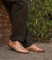 A person wearing Bed Stu Anchiano oxford-style lace-up brown dress shoes and dark pants stands on gravel by a brick wall and green foliage.