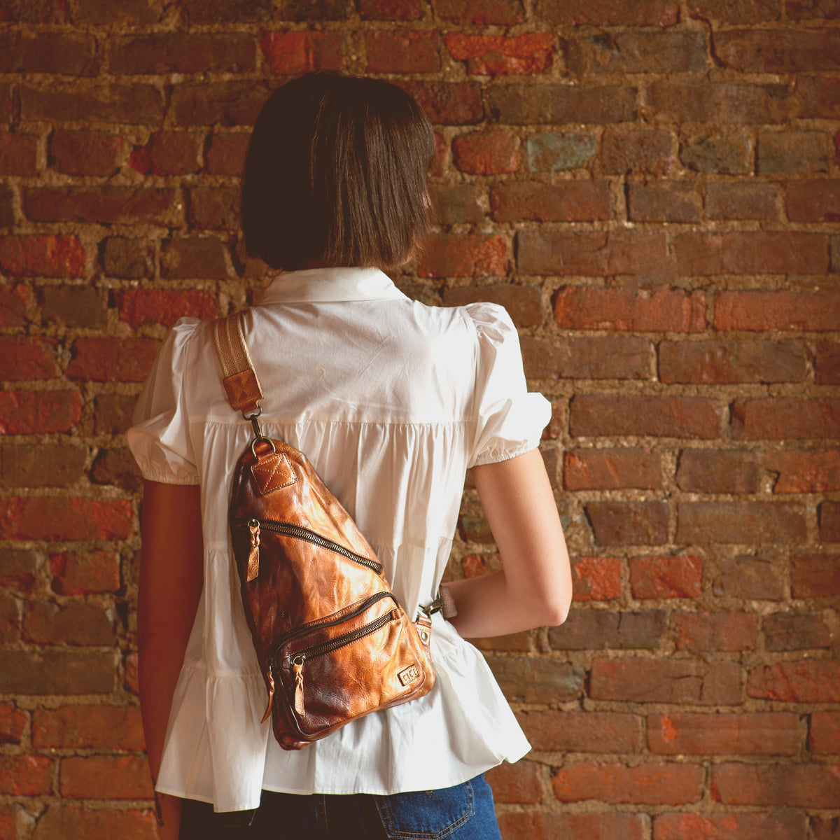 A practical woman with the versatile Bed Stu Andie leather sling bag poses in front of a brick wall.
