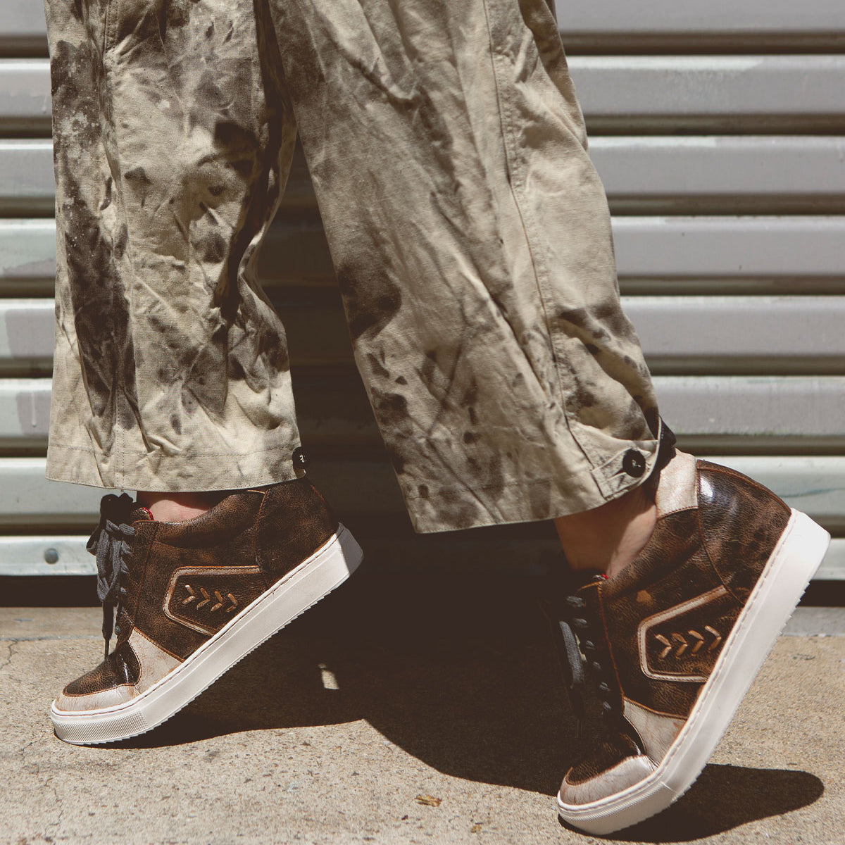 A person wearing Bed Stu Andromeda brown high-top sneakers with a street-smart design, paired with tan, paint-splattered pants, stands on a concrete floor in front of a corrugated metal wall.