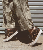 A person wearing Bed Stu Andromeda brown high-top sneakers with a street-smart design, paired with tan, paint-splattered pants, stands on a concrete floor in front of a corrugated metal wall.