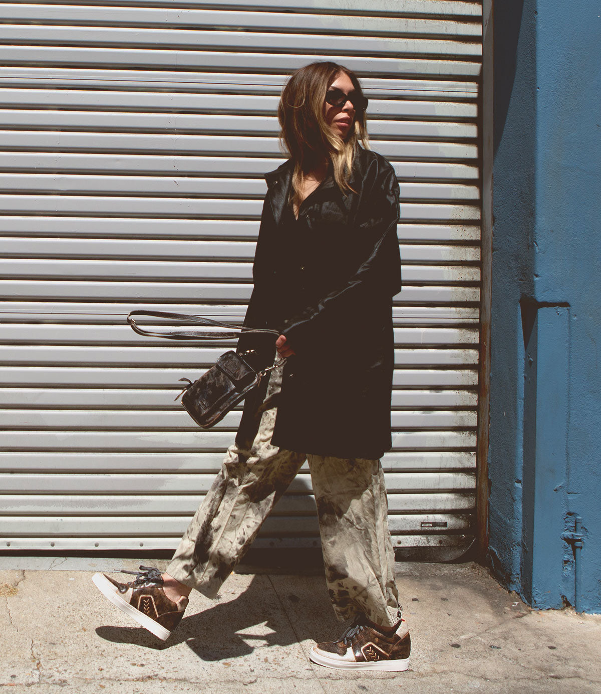A person wearing Andromeda sunglasses by Bed Stu, a black coat, patterned pants, and high-top sneakers with a hidden heel is walking on a sunlit sidewalk in front of a metal shutter.