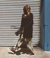 A person wearing Andromeda sunglasses by Bed Stu, a black coat, patterned pants, and high-top sneakers with a hidden heel is walking on a sunlit sidewalk in front of a metal shutter.