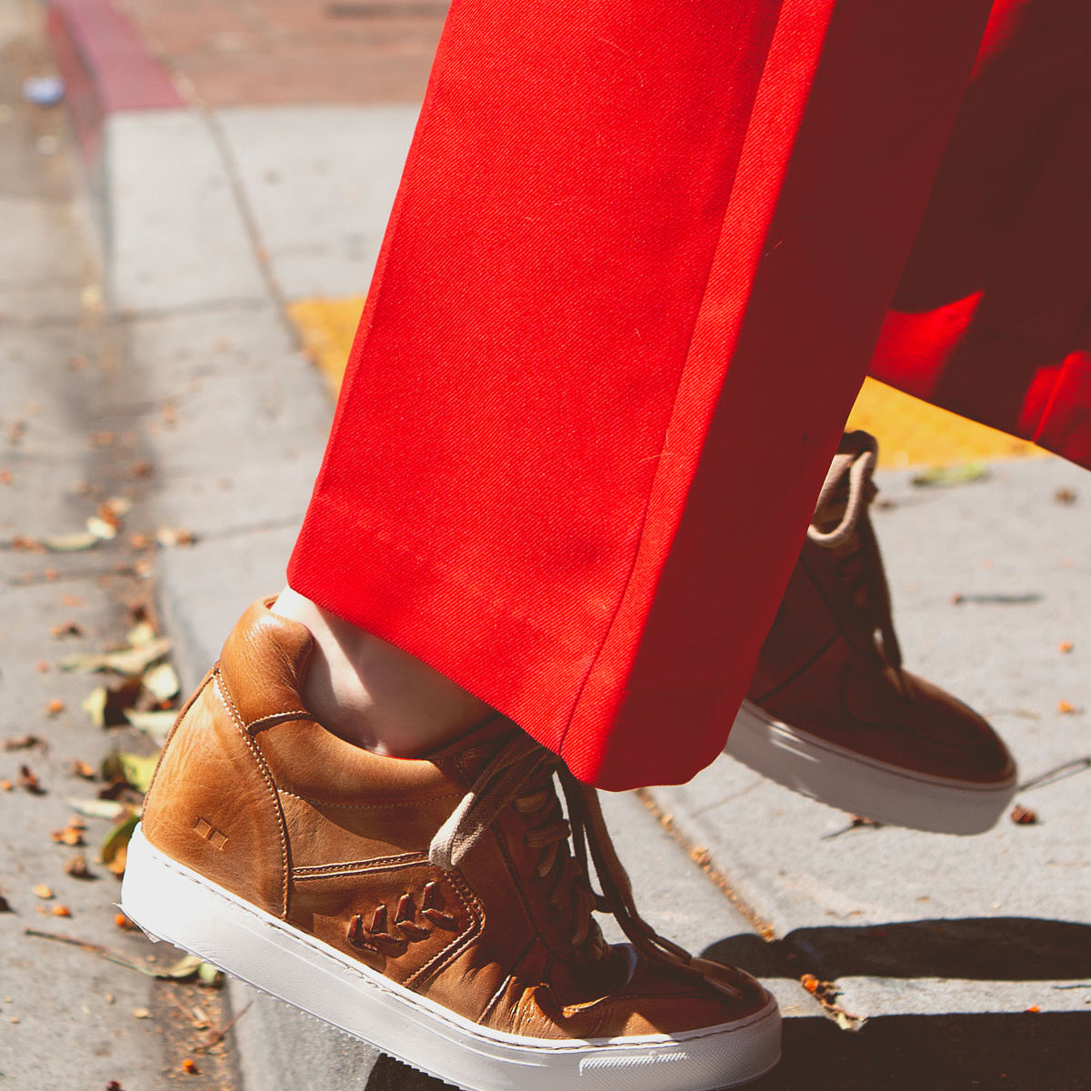 A person wearing bright red pants and the Bed Stu Andromeda tan leather high-top sneakers with a street-smart design is stepping off a curb onto the street.