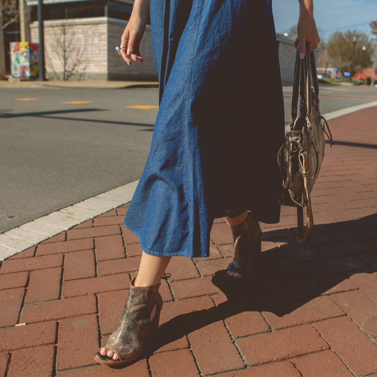 A person wearing a blue dress and Bed Stu Angelique II ankle boots with back lacing, carrying a handbag while walking across a brick-paved area.