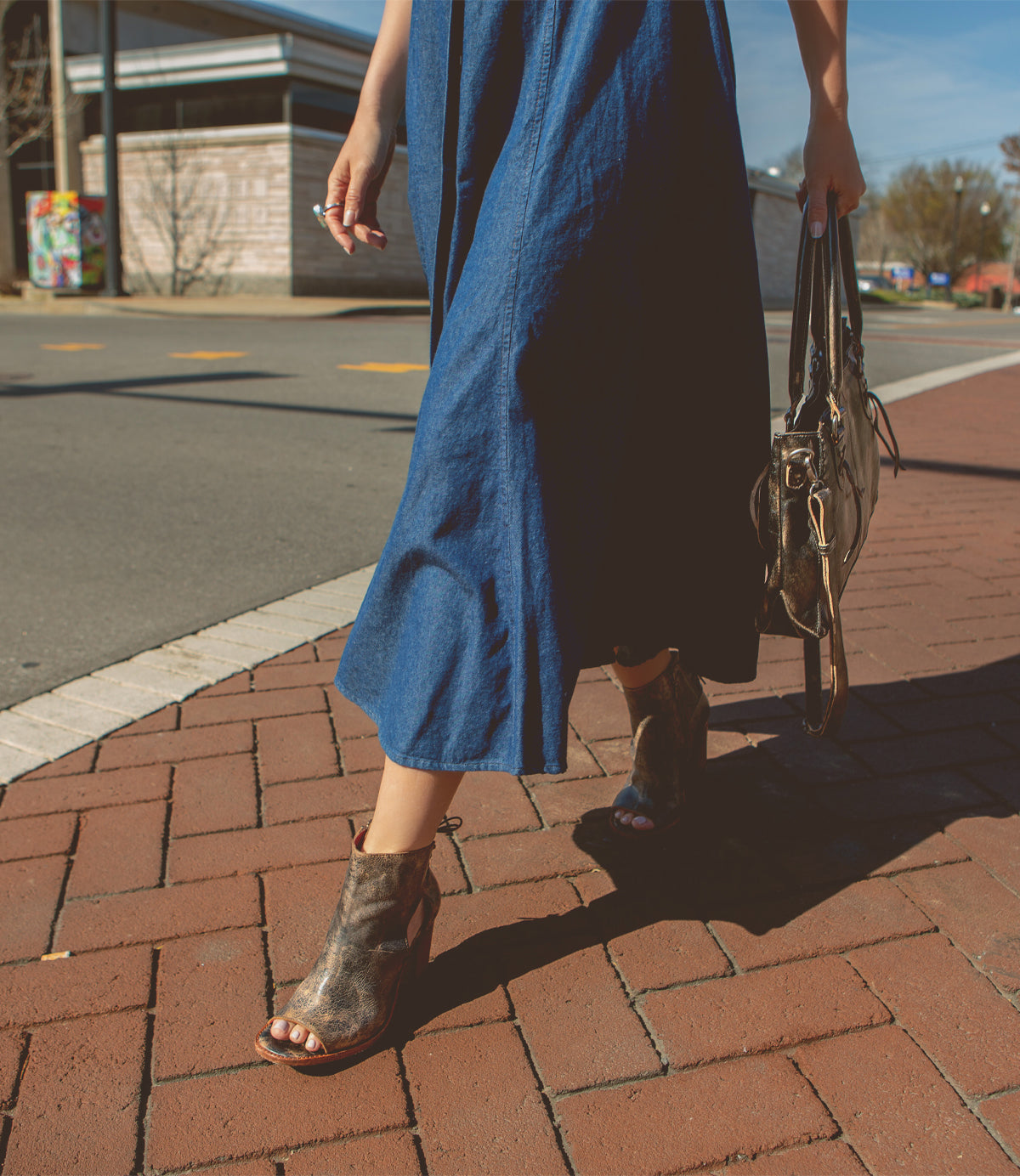 A person wearing a blue dress and Bed Stu Angelique II ankle boots with back lacing, carrying a handbag while walking across a brick-paved area.
