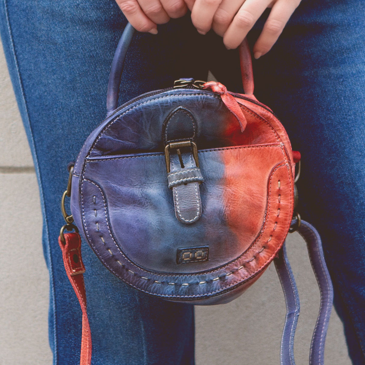 A person holds a small round leather handbag with a worn, multicolored design featuring a buckle and an adjustable crossbody strap. They are wearing blue jeans and carrying the Arenfield by Bed Stu.