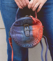 A person holds a small round leather handbag with a worn, multicolored design featuring a buckle and an adjustable crossbody strap. They are wearing blue jeans and carrying the Arenfield by Bed Stu.