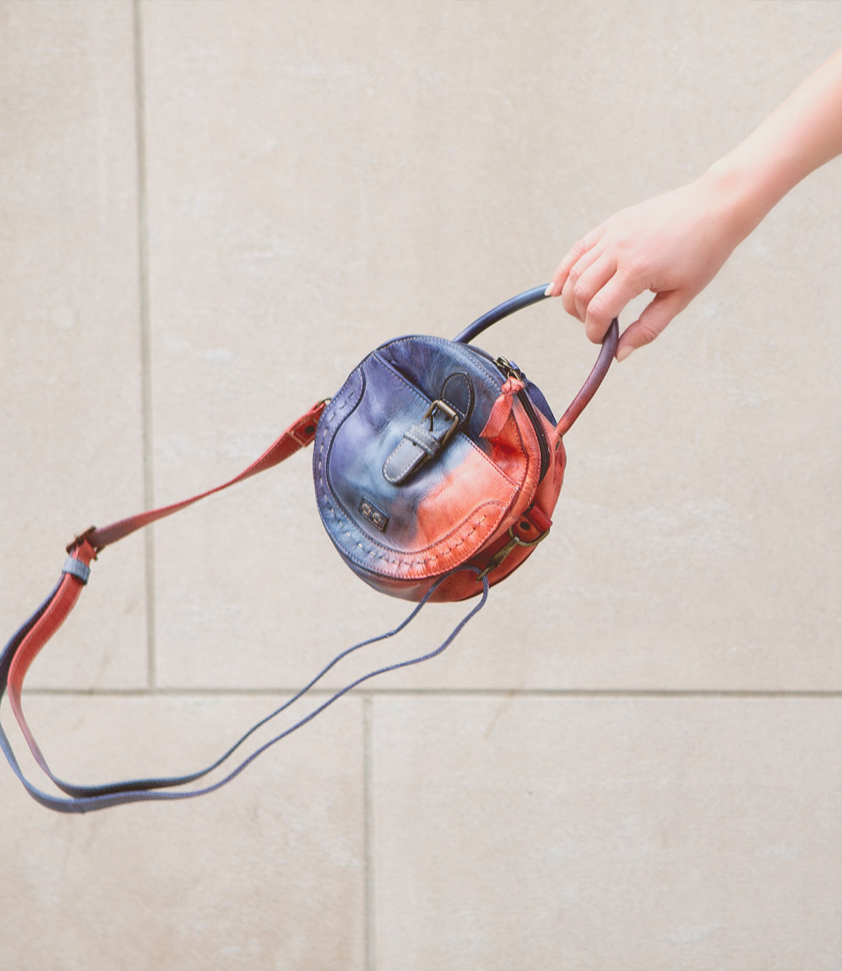 A hand holds a unique round compact crossbody bag, Arenfield by Bed Stu, with a blue and red gradient design, featuring an adjustable crossbody strap, against a beige background.