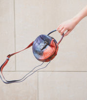 A hand holds a unique round compact crossbody bag, Arenfield by Bed Stu, with a blue and red gradient design, featuring an adjustable crossbody strap, against a beige background.
