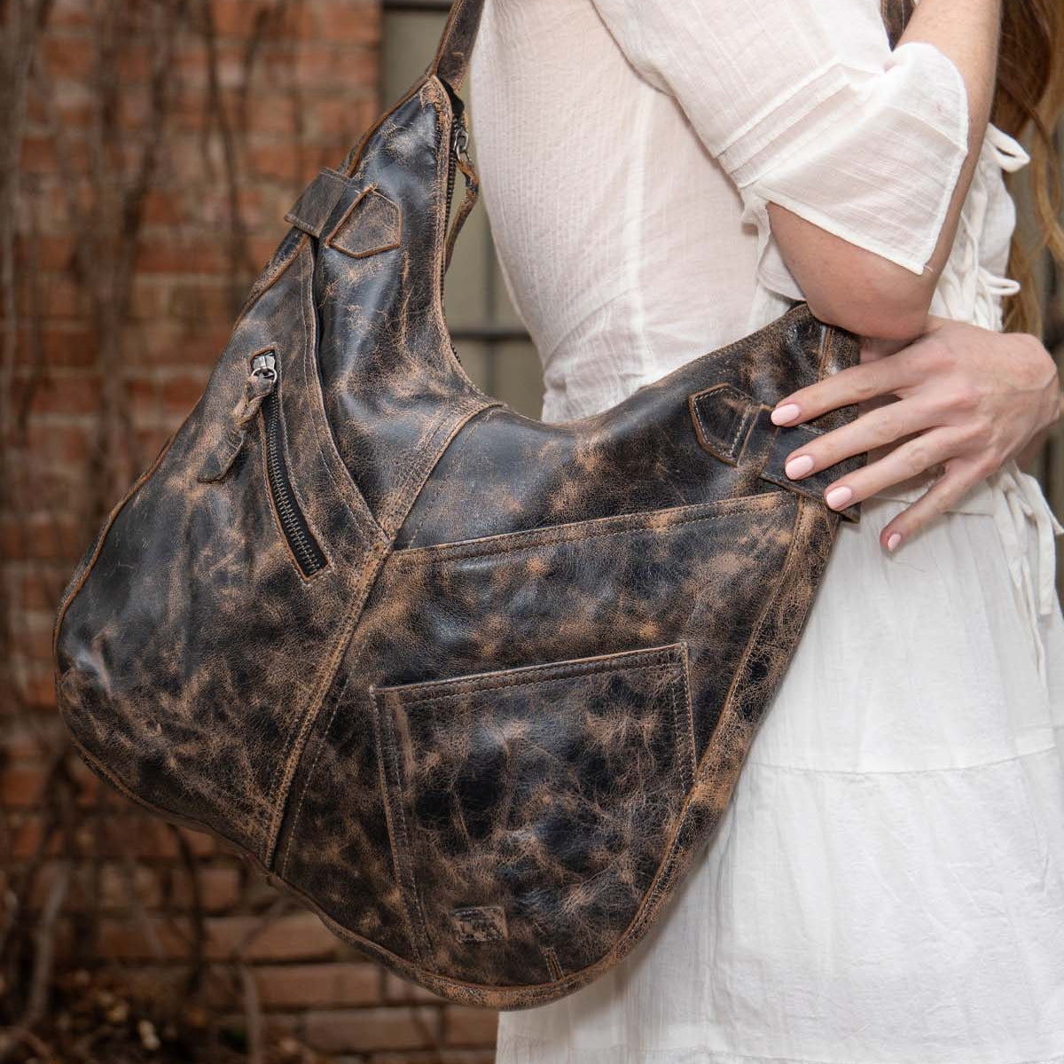 A person in a white dress carries the Bed Stu Argus, a large handcrafted leather shoulder bag with multiple front pockets, while standing outdoors near a brick wall.