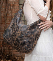 A person in a white dress carries the Bed Stu Argus, a large handcrafted leather shoulder bag with multiple front pockets, while standing outdoors near a brick wall.