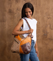 A woman in a white sleeveless top and jeans holds the Bed Stu Argus handcrafted leather shoulder bag, featuring oversized front pockets, while standing before a wooden backdrop.