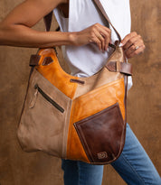 A person in a white sleeveless top holds the Bed Stu Argus, a handcrafted leather shoulder bag featuring tan, orange, and dark brown sections with oversized front pockets.