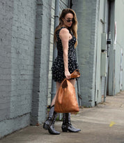 A woman in a black patterned dress, sunglasses, and cowboy boots carries the Bed Stu Ariel EM limited-edition tote bag as she walks along an urban sidewalk.