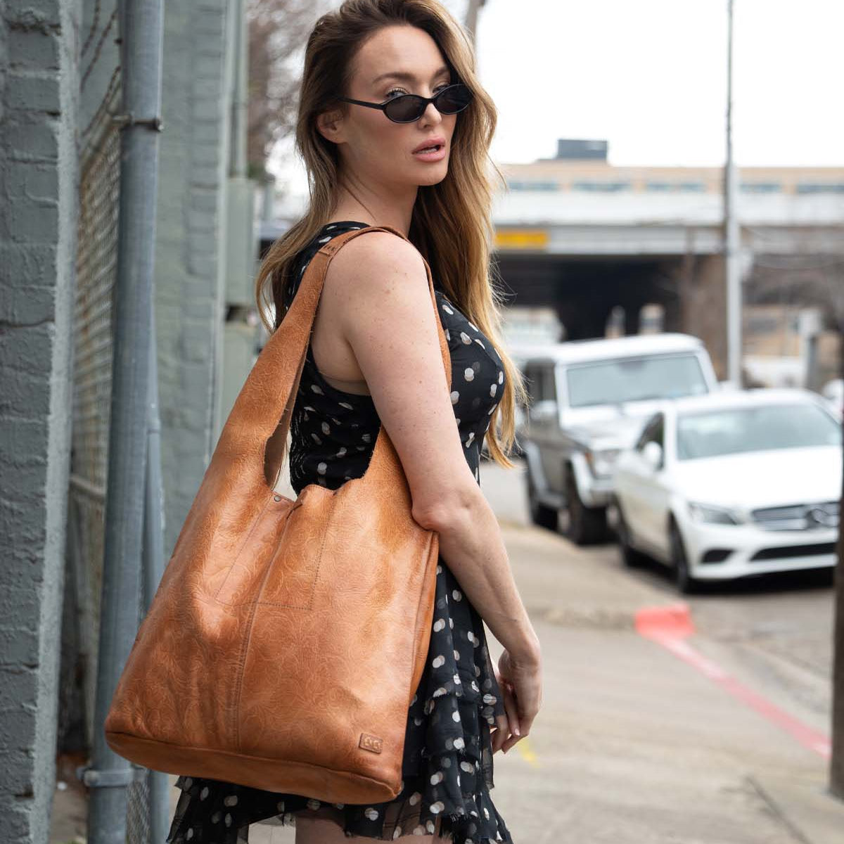 A woman in a black polka dot dress and sunglasses stands on a sidewalk, carrying the Bed Stu Ariel EM limited-edition embossed tan leather tote bag over her shoulder.
