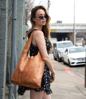 A woman in a black polka dot dress and sunglasses stands on a sidewalk, carrying the Bed Stu Ariel EM limited-edition embossed tan leather tote bag over her shoulder.