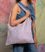 A woman in a sleeveless denim outfit carries the Bed Stu Ariel II tote, known for its spacious compartment, over her shoulder against a pastel tie-dye background.
