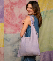 A woman in a sleeveless denim outfit holds the Bed Stu Ariel II, a large, lightweight tote with a spacious compartment, standing before a pastel tie-dye background.
