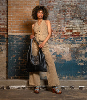 A woman stands against a brick wall, wearing a tan vest and pants set, blue platform sandals, and carrying the Bed Stu Ariel II tote with a spacious main compartment.