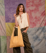 A woman stands before a vibrant patchwork backdrop, wearing a cream top and dark pants, carrying the spacious yellow Ariel II tote bag by Bed Stu over her shoulder.