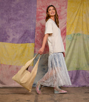 A woman smiles while walking, wearing a beige oversized T-shirt, a sheer lace skirt, pastel boots, and carrying the Bed Stu Ariel II tote with a spacious compartment. Colorful fabric panels hang in the background.