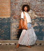 A person stands before a textured brick wall, wearing a white t-shirt, sheer lace skirt, brown sandals, and holding the Bed Stu Ariel II tote—a minimalism-inspired lightweight bag with a spacious main compartment.